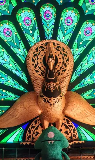 Keltar sitting small in front of a carved orange peacock sculpture with a fan of green and teal stained-glass feathers.