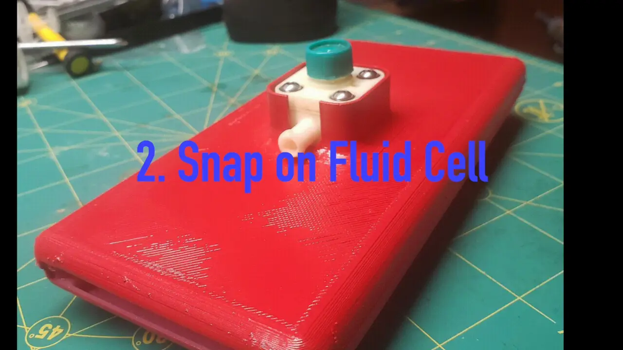 A red handheld device with a white fluid cell on a green cutting mat, under the words 2. Snap on Fluid Cell